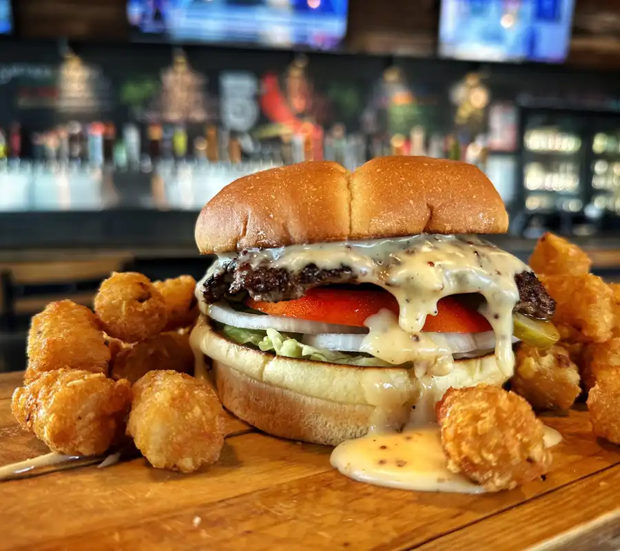 smash burger with dripping beer cheese tomato onion lettuce on a bun surrounded with tater tots